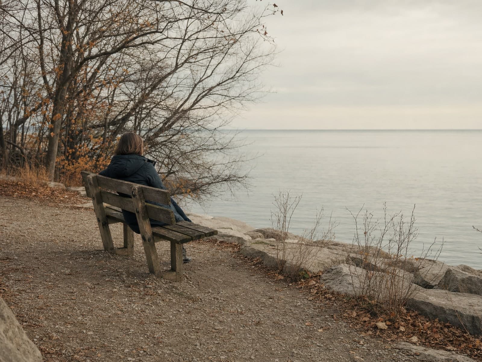 A gravel path along a lake shore in late autumn, a wooden bench turned toward flat pale water