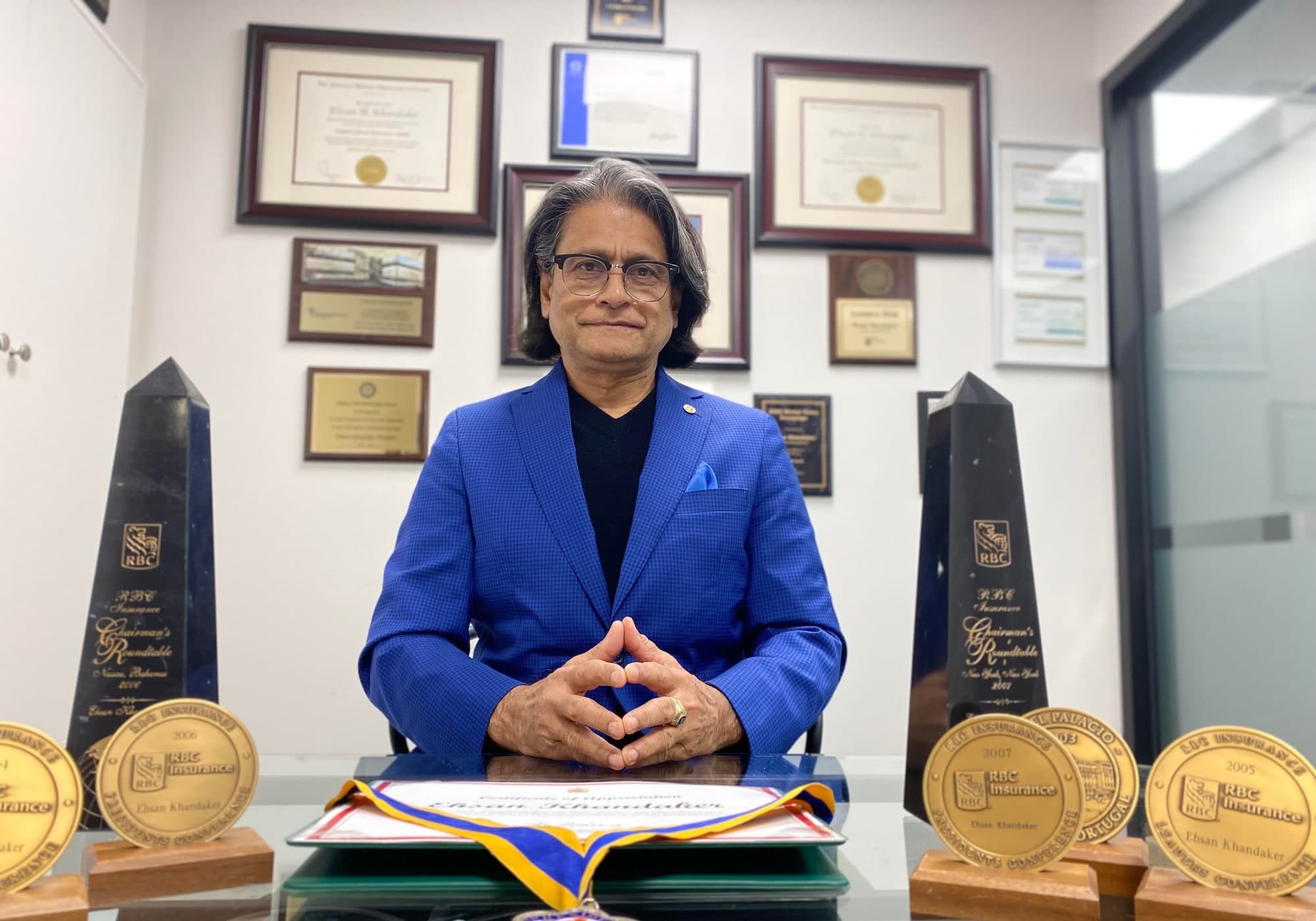 Ehsan Khandaker at his desk, between two RBC Insurance Chairman's Roundtable awards, with framed designations behind him