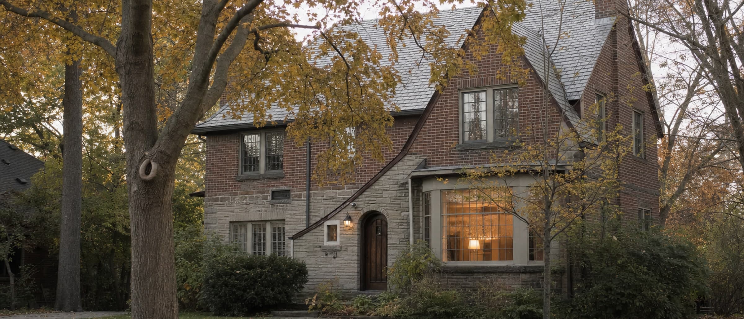 A brick and stone family house across a lawn, its maples turning, one window lit