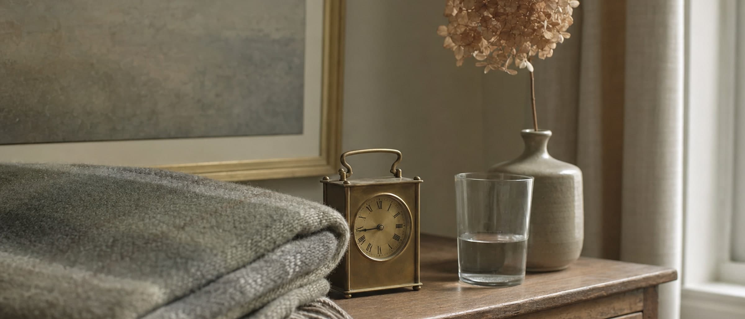 A folded wool blanket, a small brass clock and a dried hydrangea on a sideboard